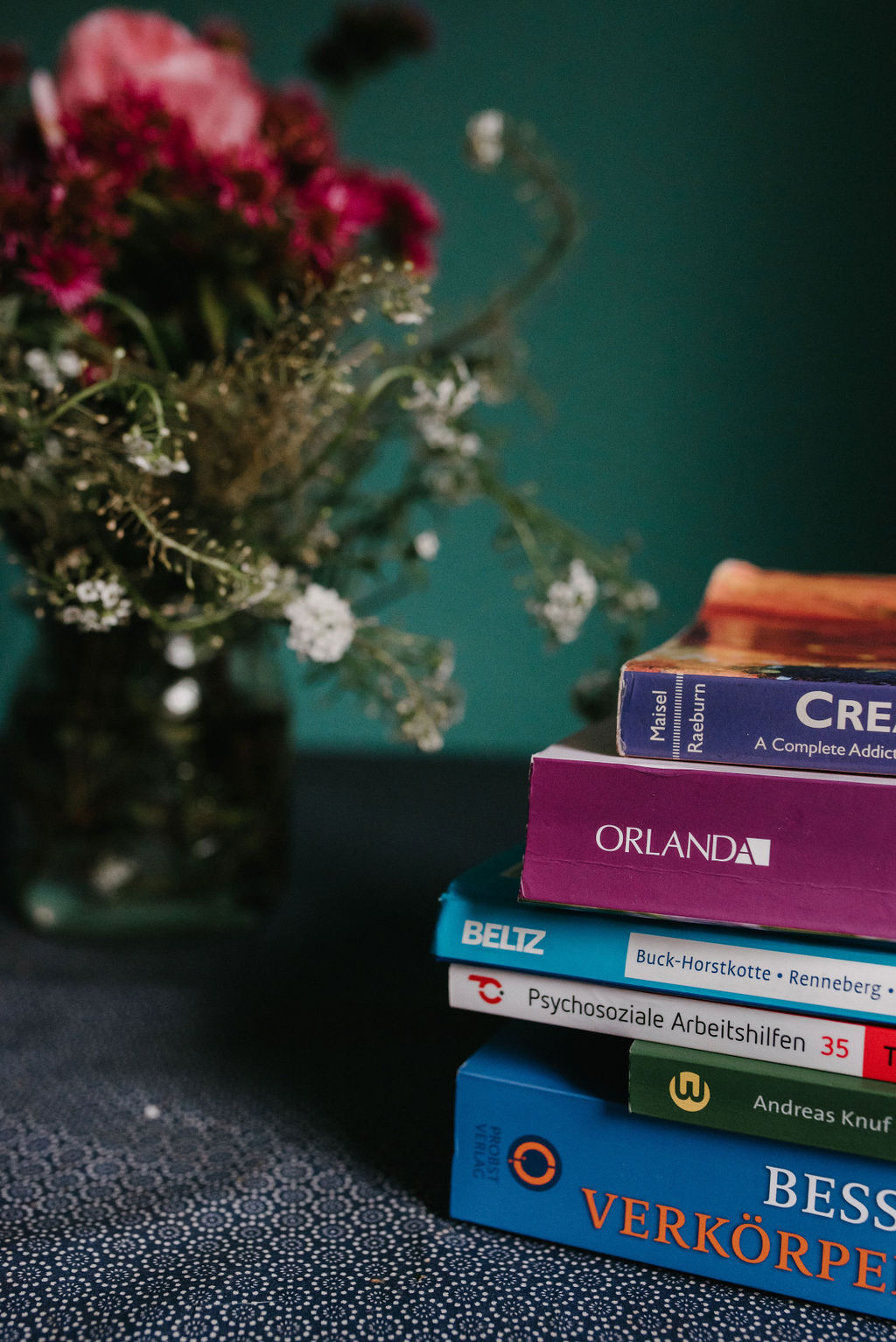 Ein Stapel mit Büchern zu psychosozialen Themen liegt auf einem Tisch, dahinter steht ein Blumenstrauß