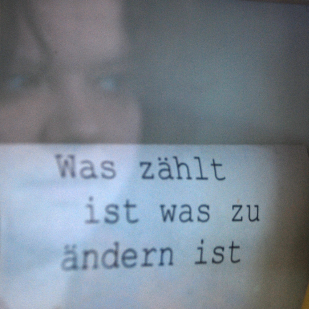 Im Hintergrund die Spiegelung von Natalies Gesicht in einer Glasscheibe. Im Vordergrund ein Zettel, auf dem steht: "Was zählt, ist was zu ändern ist."