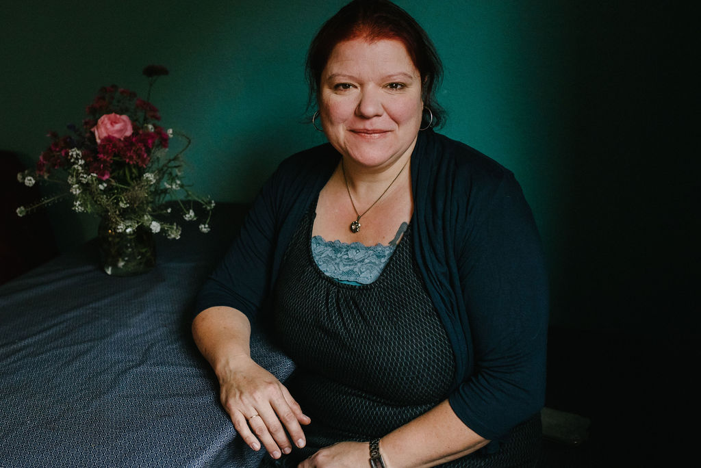 Natalie sitzt am Küchentisch vor einer türkisen Wand und schaut etwas angespannt in die Kamera.